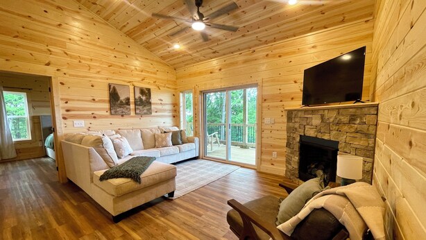 A cozy cabin interior with a wooden interior, a stone fireplace, a flat-screen TV, and a sliding glass door leading to a deck. - Beautiful Mountain View - Charming 2-bedroom house in Murphy, NC (Murphy)