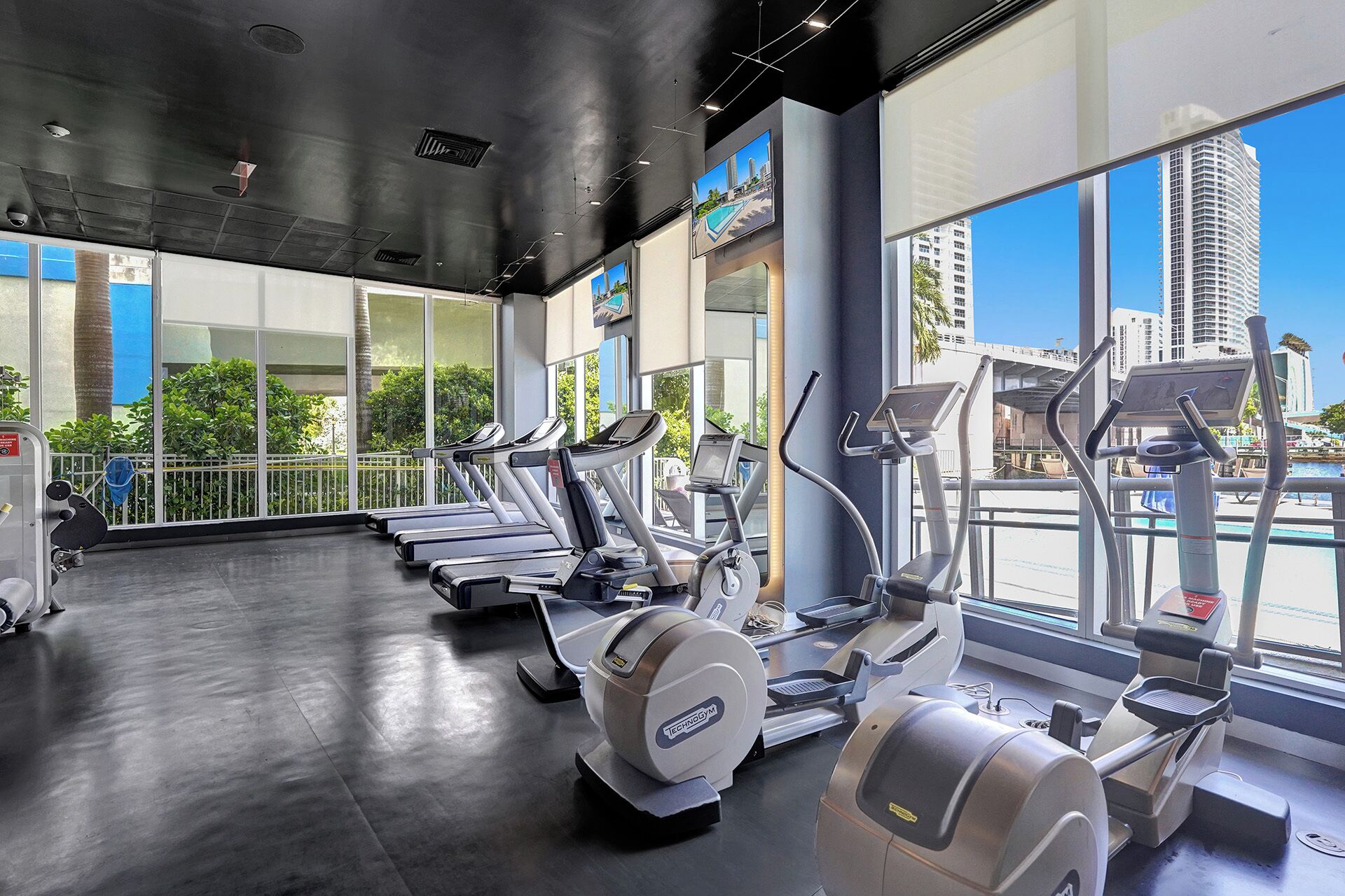 A gym with multiple treadmills and elliptical machines, large windows with a city view, and a polished concrete floor.