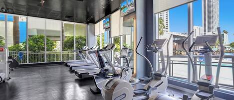 A gym with multiple treadmills and elliptical machines, large windows with a city view, and a polished concrete floor.