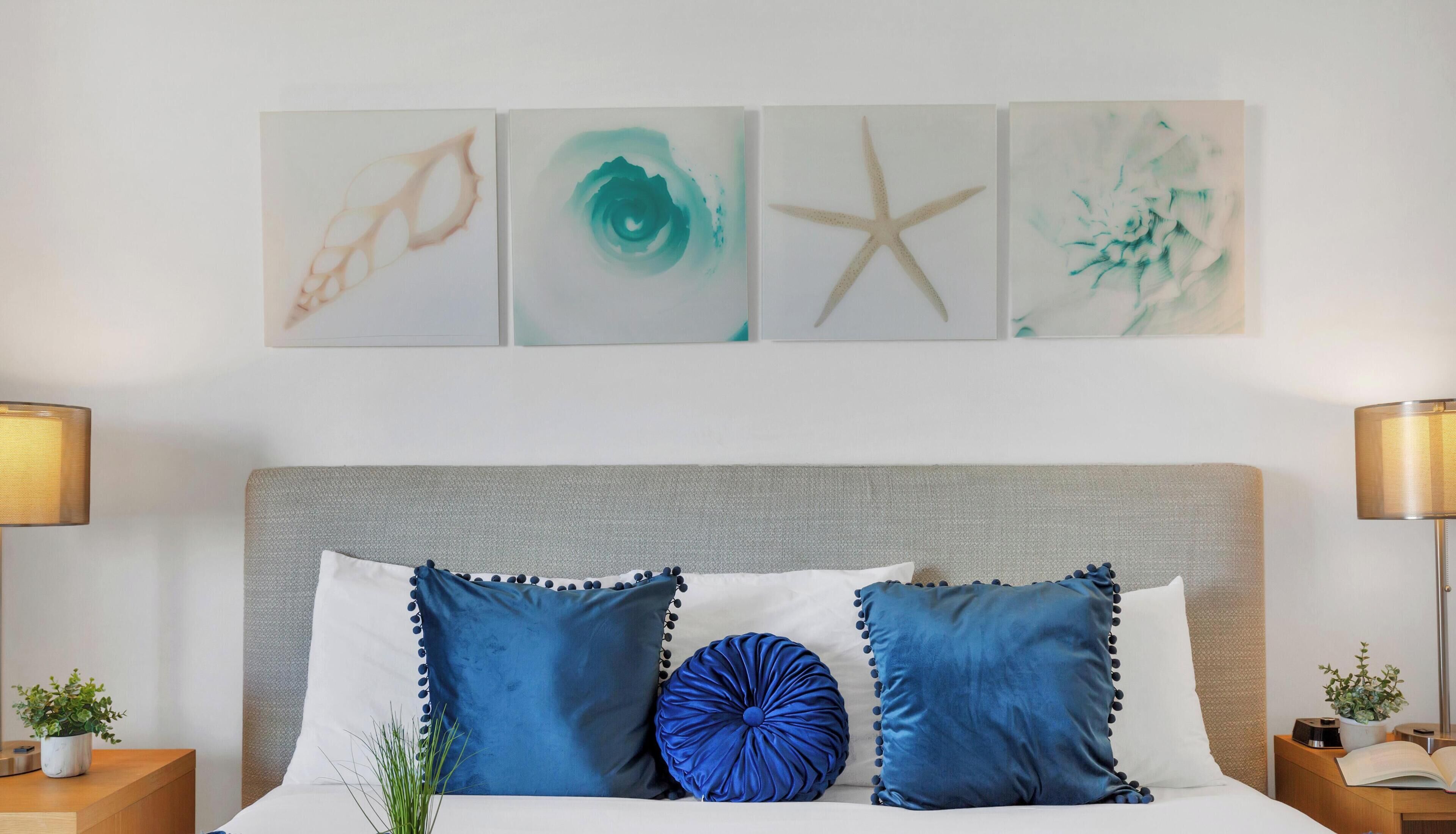 A bed with blue and white pillows, a grey headboard, and four wall art pieces above.