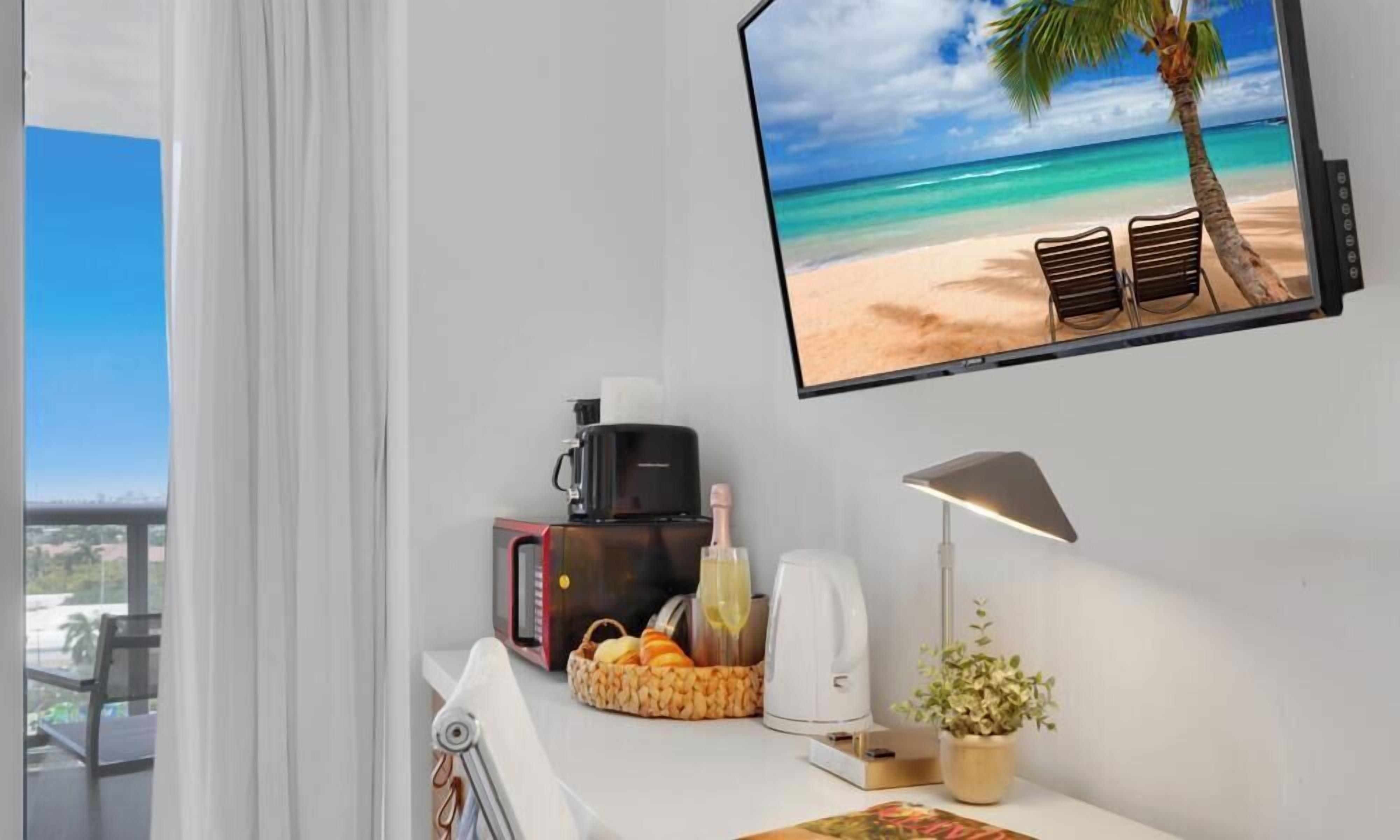 Ett hotellrum med ett stort fönster, en platt-TV som visar en strandscen, en kaffebryggare, en brödrost, en korg med frukt och en liten krukväxt.