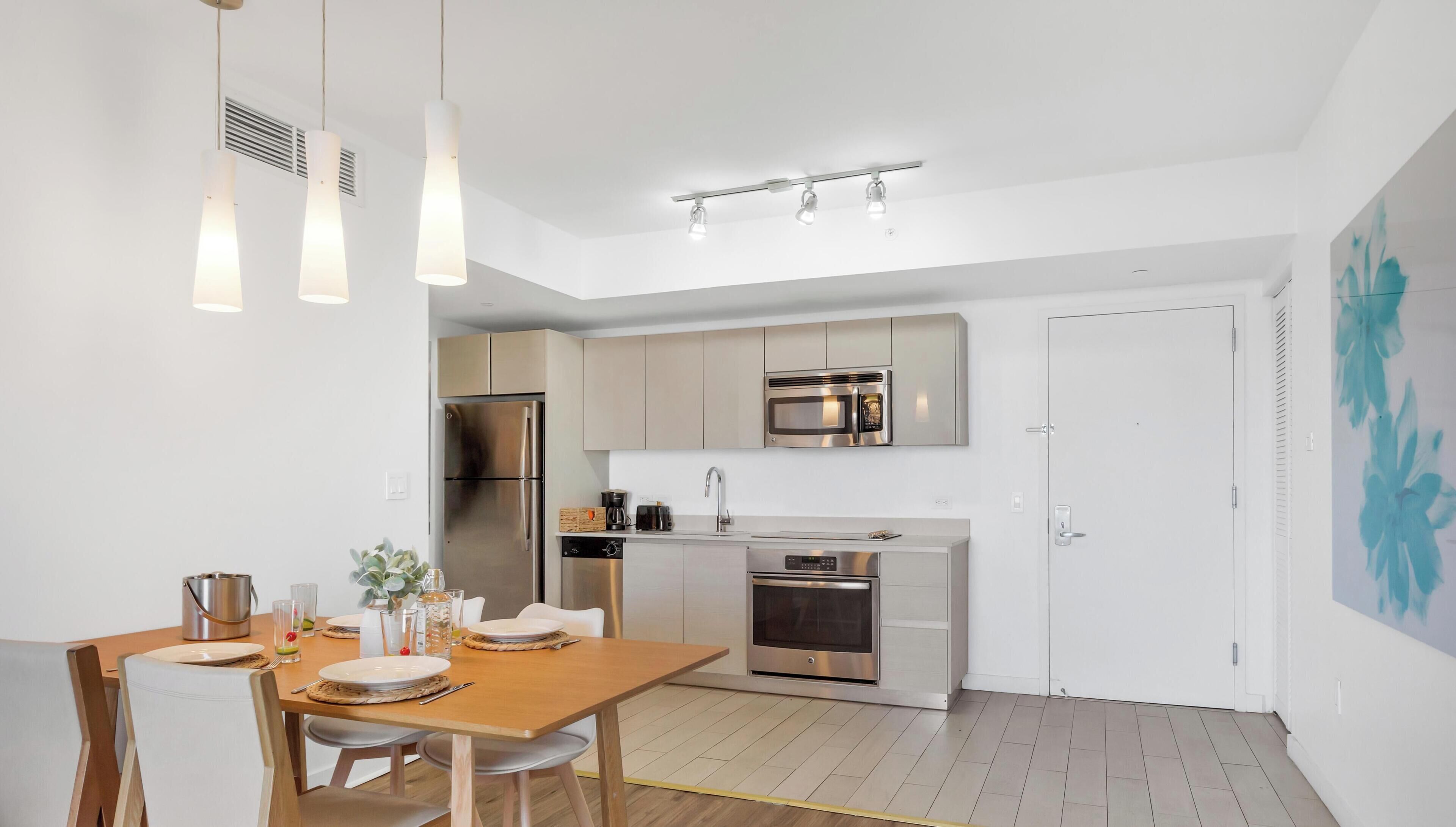 Una cocina moderna con electrodomésticos de acero inoxidable, una zona de comedor con mesa y sillas de madera, e iluminación colgante.