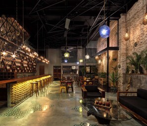 Un espace bar avec un mur en briques, des étagères en bois et des luminaires suspendus. Des tables et des chaises sont disposées pour accueillir les clients.