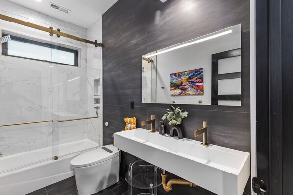 Une salle de bain moderne avec une douche à l’italienne en verre, un meuble-lavabo blanc avec un robinet doré et un grand miroir.