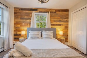 Un dormitorio con panel de pared de madera, un ventanal con cortinas, una cama con ropa de cama blanca, dos lámparas de noche y una puerta.