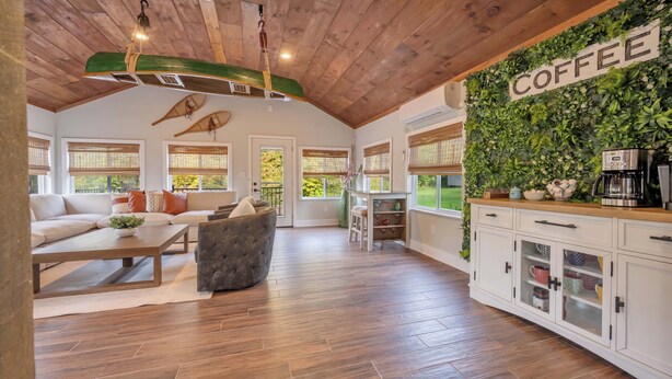 A spacious living area with a wooden ceiling, a sofa, a coffee station, and a wall decorated with greenery. - Heated Pool • Hot Tub• Game Room• Fire Pit• 6 Bdrms (Forest City)