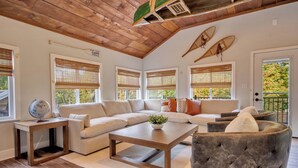 A spacious living room with a wooden ceiling, large windows with bamboo blinds, a beige sofa, a coffee table, and a grey armchair. - Heated Pool • Hot Tub• Game Room• Fire Pit• 6 Bdrms (Forest City)