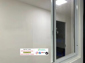 Quarto de hotel com uma janela grande, parede branca e porta. Há um aviso no canto inferior esquerdo com texto e logotipos.