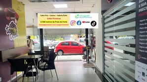 A restaurant interior with a glass partition, a red car outside, and a sign advertising 'Cabo Frio - Centro - Galeria Éxito'.