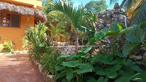 A tropical garden with a thatched-roof building, lush greenery, and a stone wall. - Suites Casa de Piedra y Colores-Sak (Tinum)