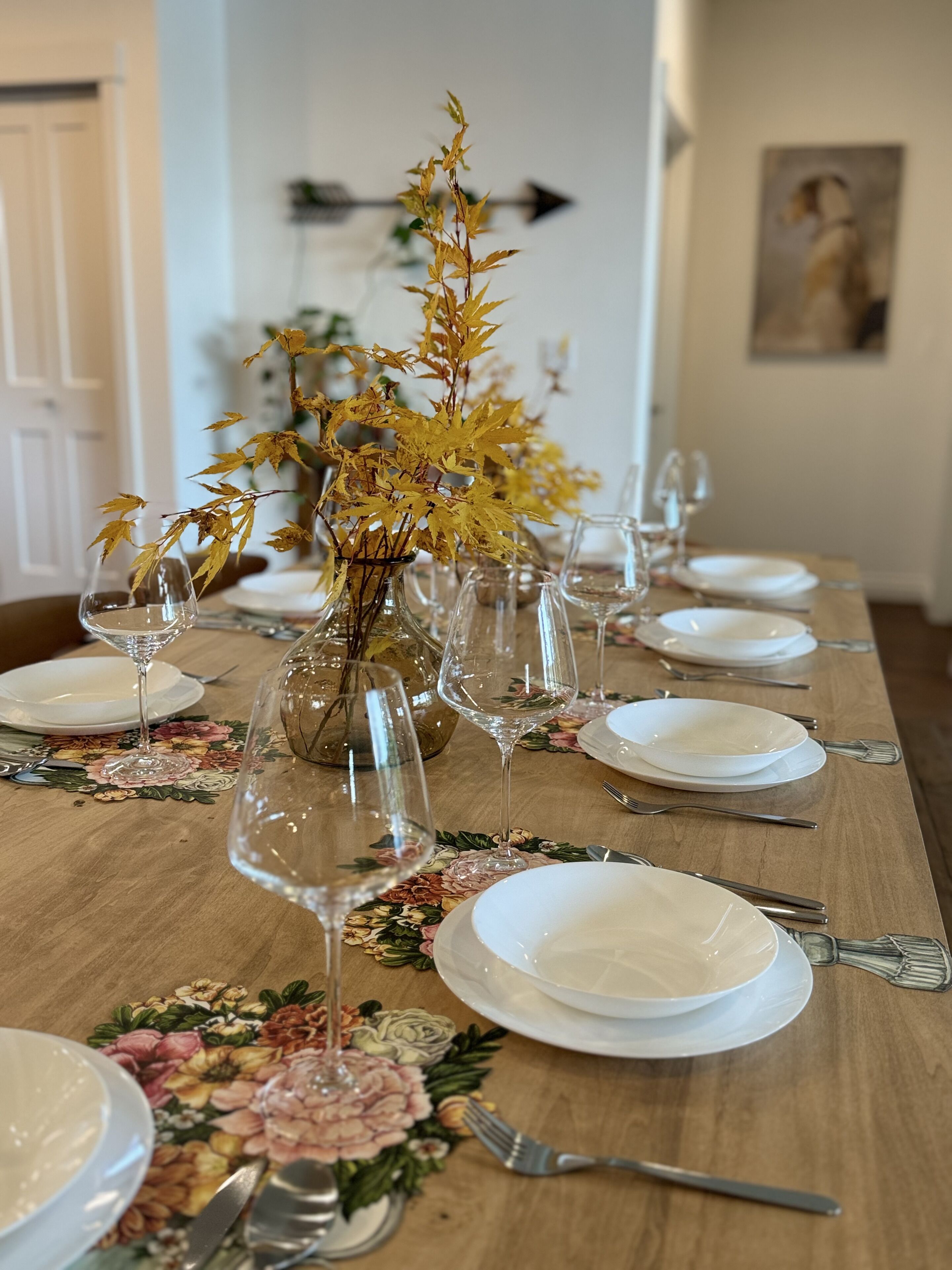 Une table à manger dressée avec de l’assiette blanche, des verres à vin transparents et un vase contenant des feuilles jaunes.