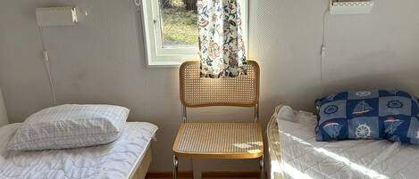 Una habitación con dos camas individuales, una silla de madera, un ventanal con cortinas florales y un equipo de aire acondicionado blanco.