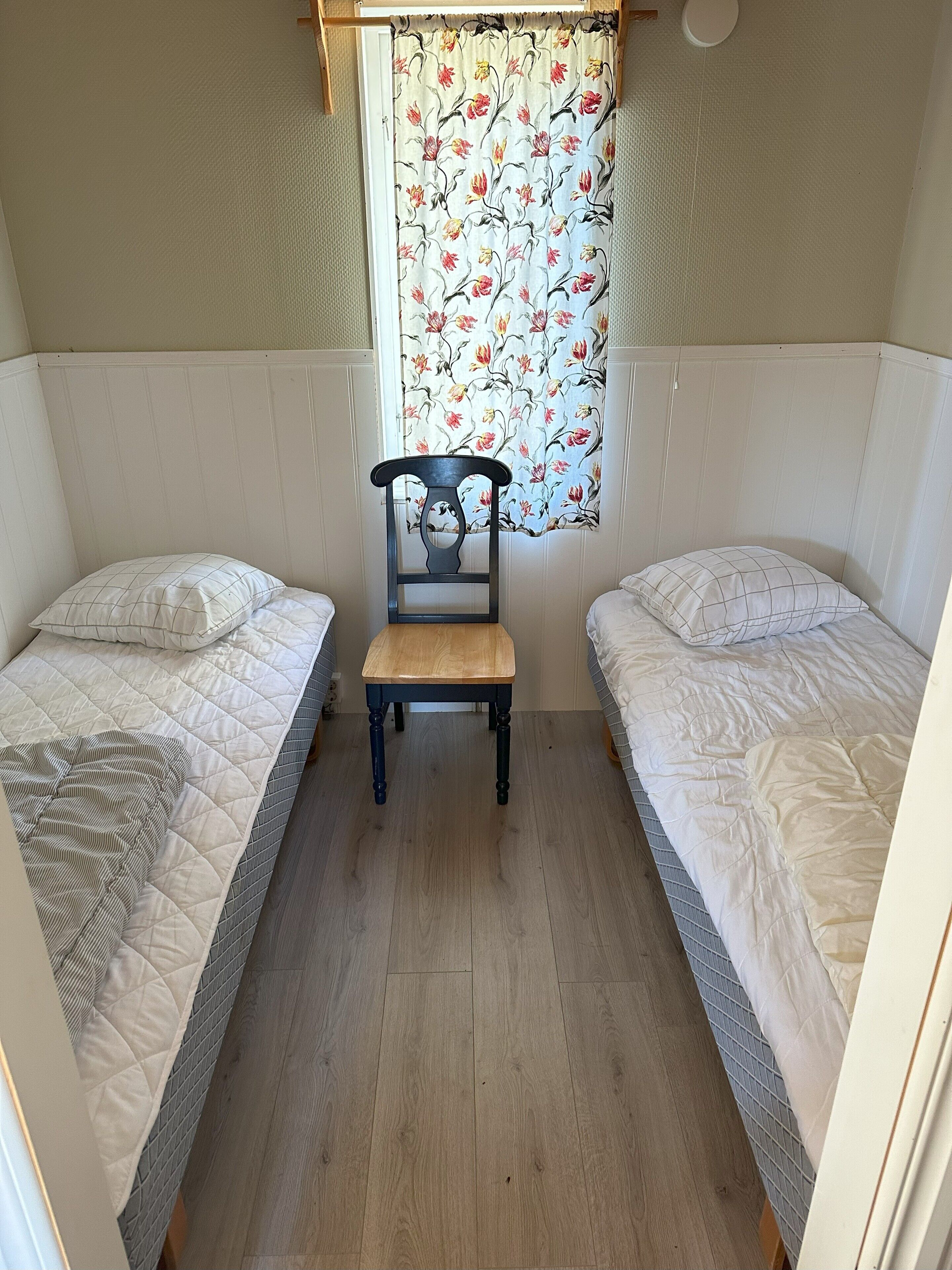 A small, cozy room with two single beds, a wooden chair, and a curtain with a floral pattern.