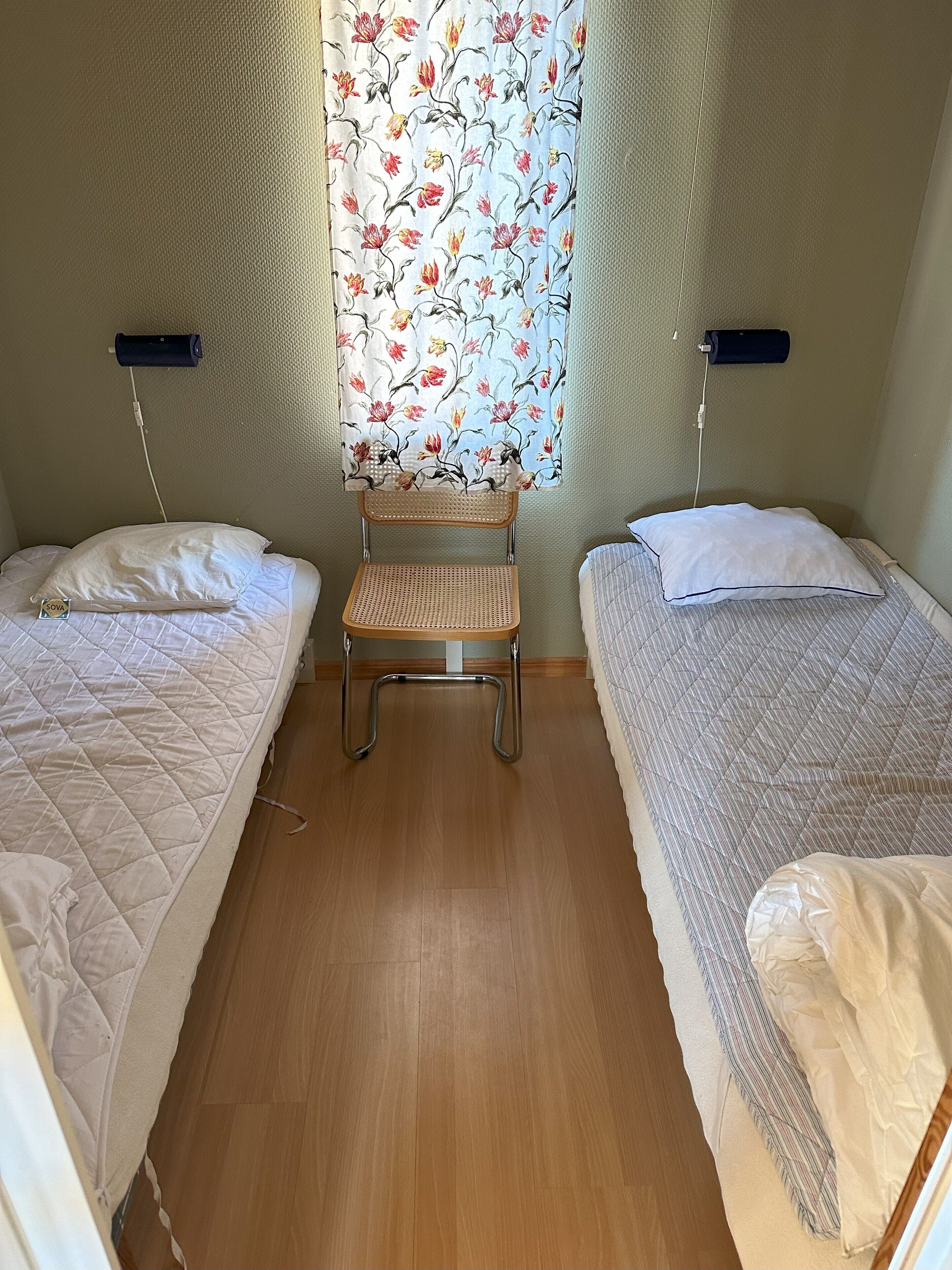 A small, cozy room with two single beds, a wooden chair, and a window with floral curtains.