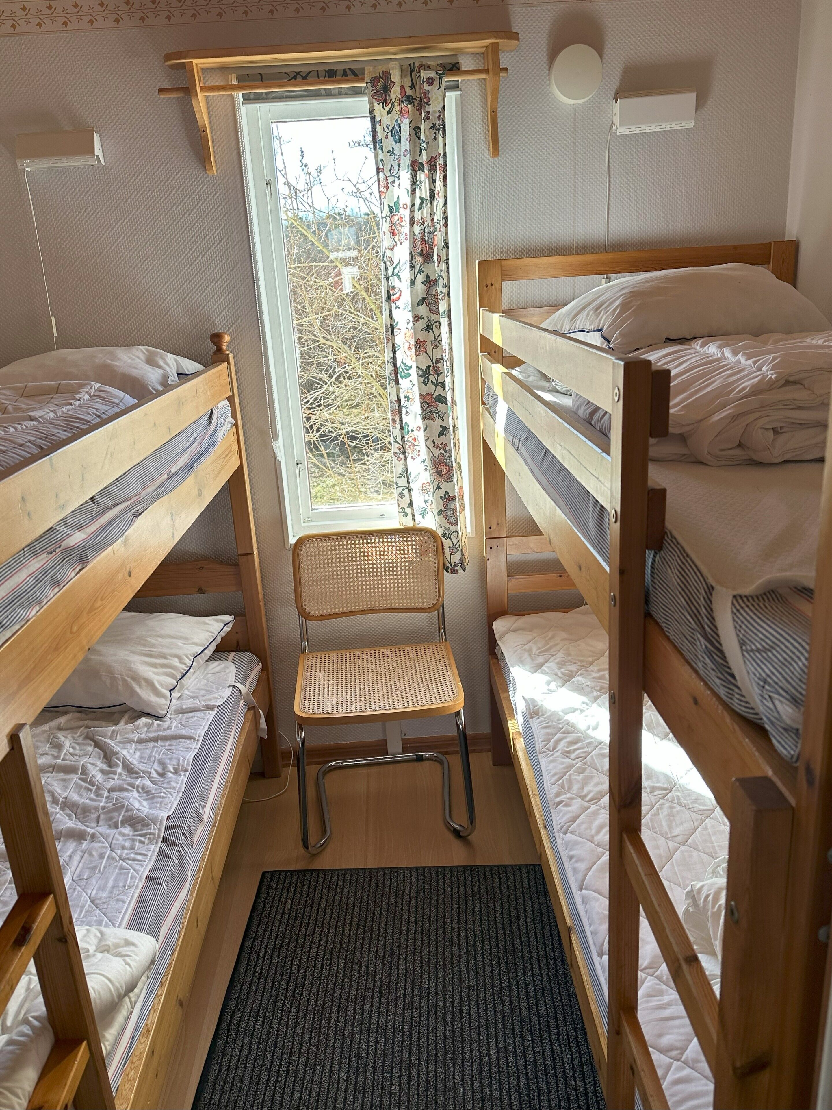 A dormitory room with bunk beds, a window with floral curtains, a standing desk, and a wall-mounted air conditioner.