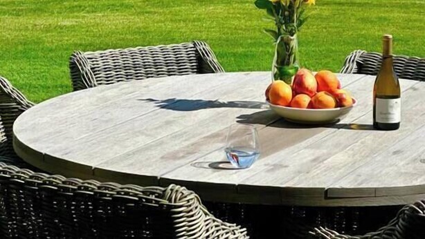 Un patio junto al lago con una mesa redonda, sillas de mimbre, una botella de vino y un cuenco de fruta.