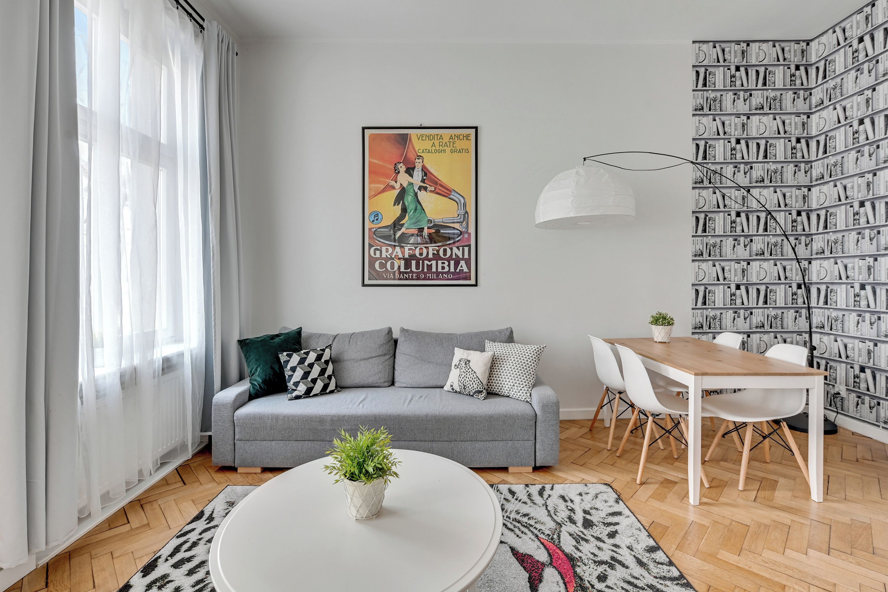 Una sala de estar moderna con un sofá gris, una mesa de centro redonda blanca con una planta, una mesa de comedor con sillas, una estantería y un póster en la pared.