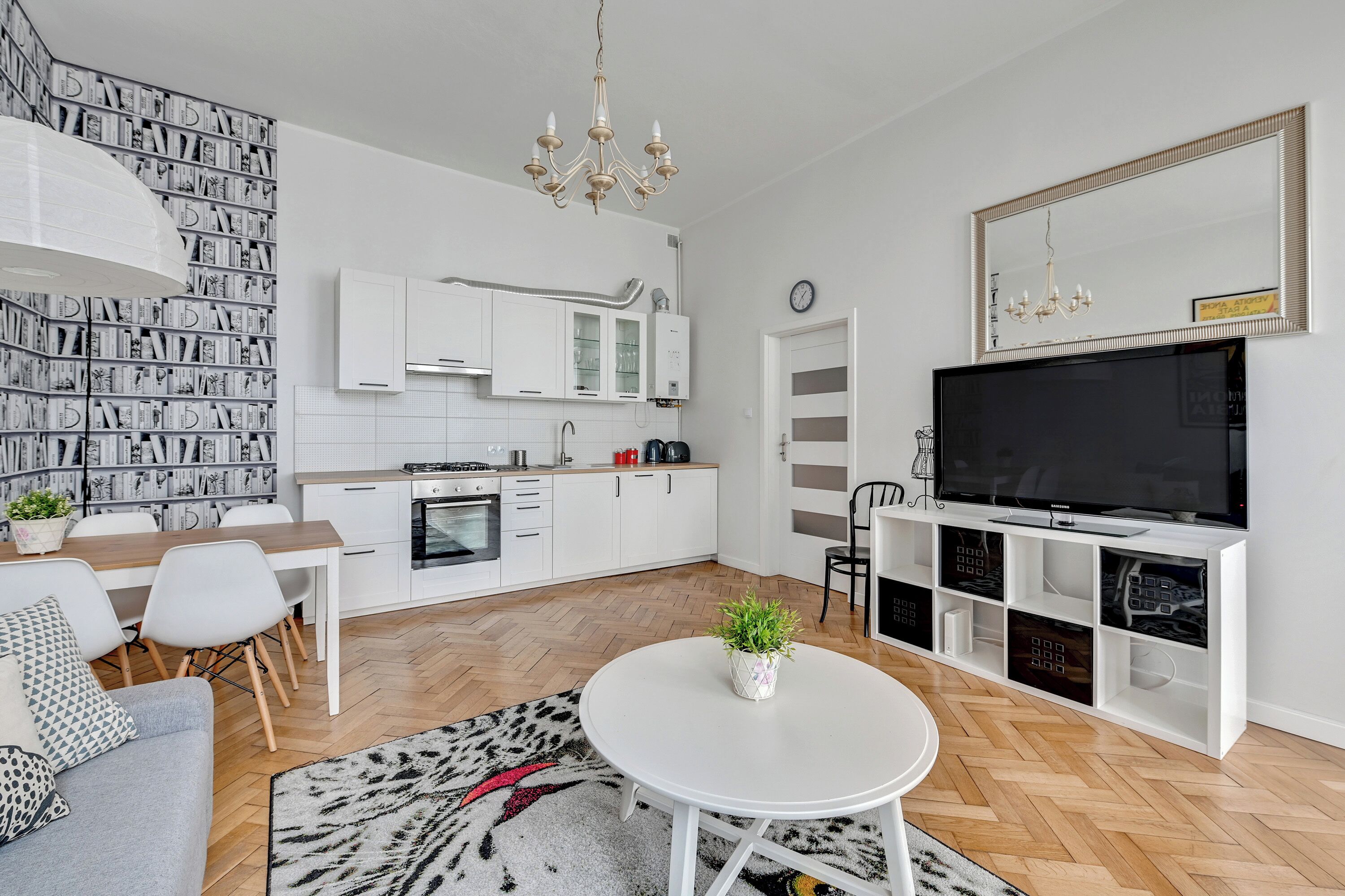 Eine moderne Küche mit wei ßen Schränken, ein Essbereich mit Tisch und Stühlen, ein Wohnzimmer mit Sofa und Fernseher sowie ein Bücherregal voller Bücher.