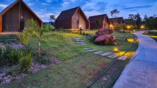 A row of modern, triangular-roofed cabins with a well-maintained lawn and pathway leading to a paved road. - Hotel Maravilha do Lago (Pedregulho)