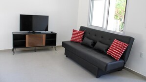 Un salon moderne avec un canapé noir, deux coussins rouges à motifs, un meuble de rangement noir intégrant un téléviseur et une fenêtre donnant sur un paysage verdoyant.