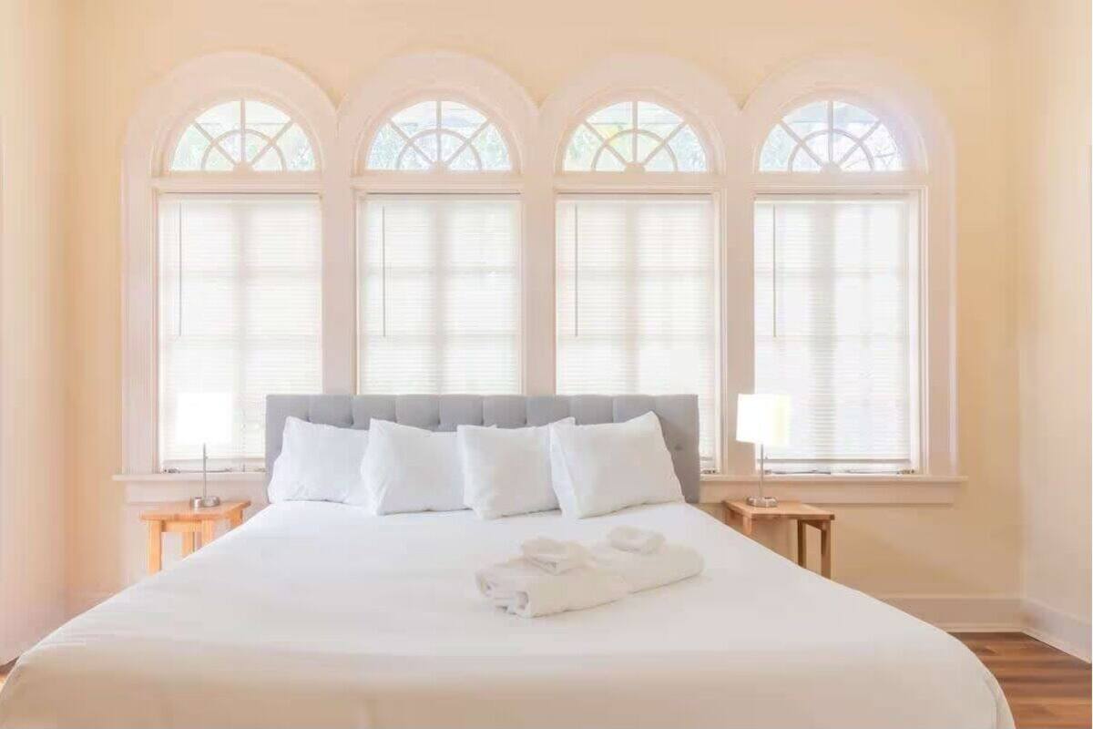 A hotel room with a large bed, white bedding, and three arched windows with blinds.