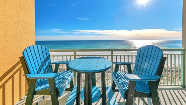 A balcony with two blue Adirondack chairs and a small round table, overlooking a beach with a clear sky and the sun shining. - 🦩Book Me Baby! Pelican Isle #604 Penthouse Paradise 🦩 (Fort Walton Beach)