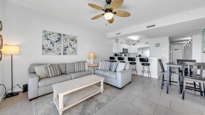 A modern living room with a grey sofa, a coffee table, and a dining area with a bar. - 🦩Book Me Baby! Pelican Isle #604 Penthouse Paradise 🦩 (Fort Walton Beach)