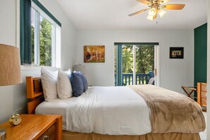 A bedroom with a bed, a nightstand, a lamp, a ceiling fan, and a view of the outdoors through a window.