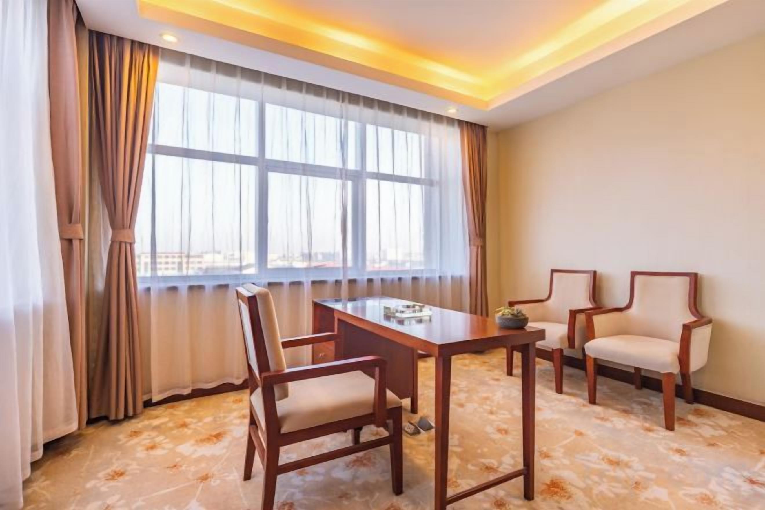 Quarto de hotel com mesa de jantar, quatro cadeiras, janela com cortinas e piso coberto de carpete.