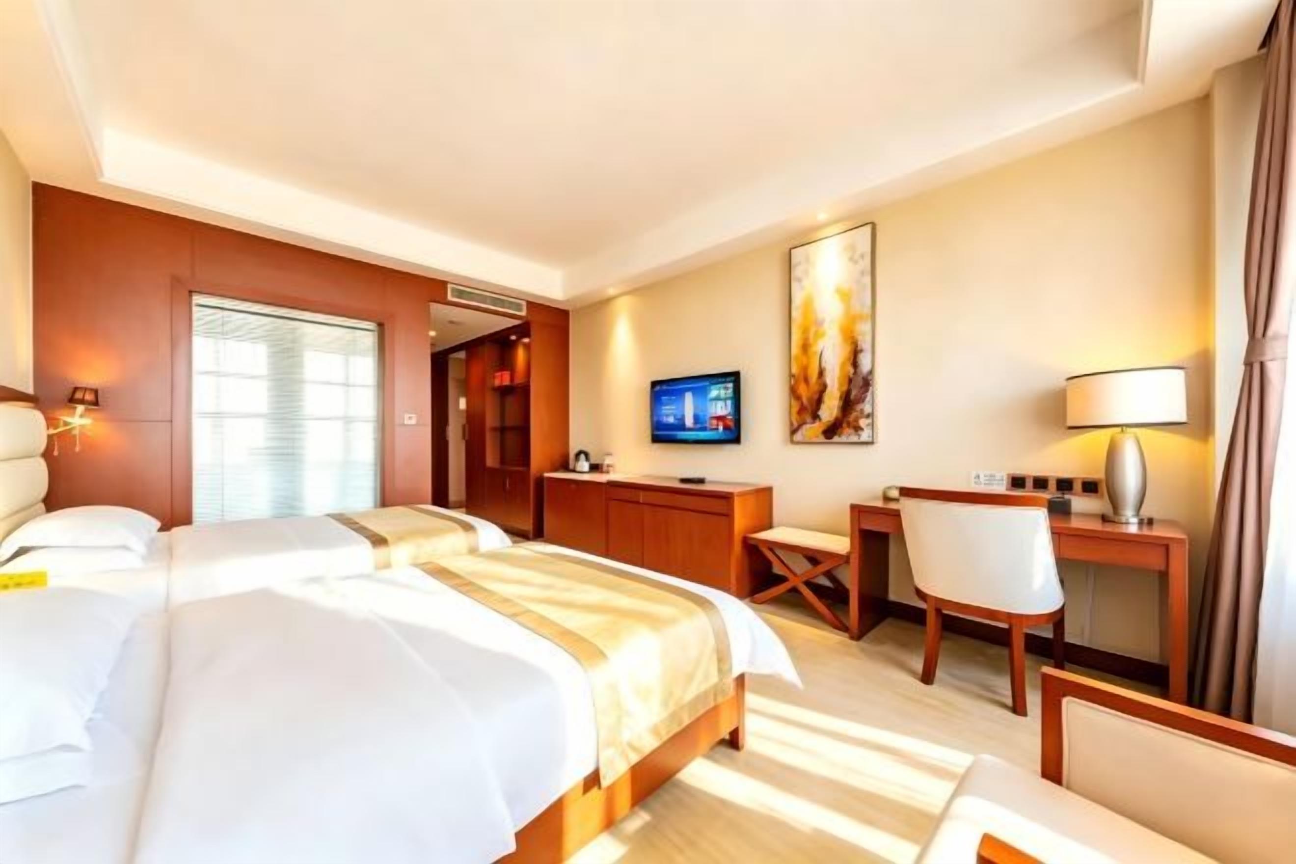A hotel room with two beds, a desk, a chair, a television, and a painting on the wall.