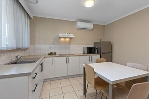 A compact kitchen with a sink, microwave, refrigerator, and a dining area with a table and chairs. - Apollo Bay Motel & Apartments (Apollo Bay)
