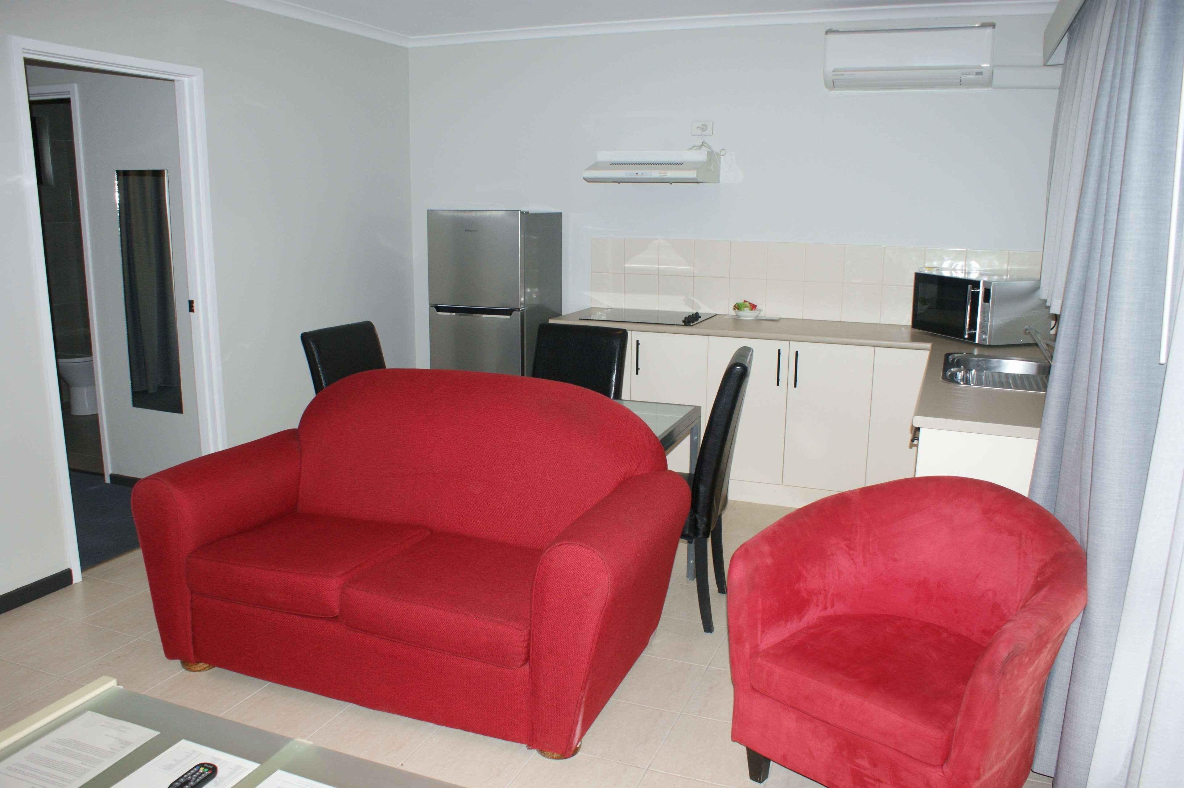 A compact kitchenette with a refrigerator, microwave, and a small dining area with a red sofa and chairs.