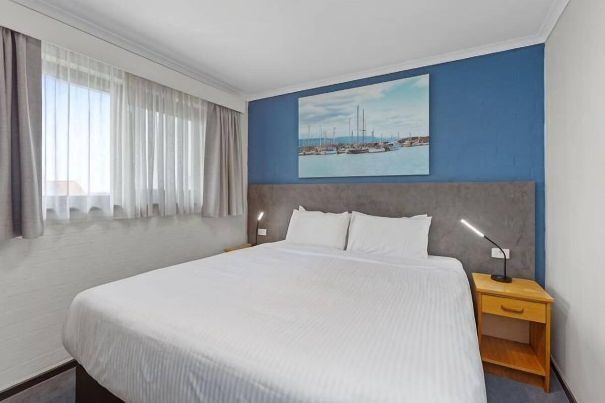 A hotel room with a large bed, a nightstand with a lamp, a painting on the wall, and a window with curtains.