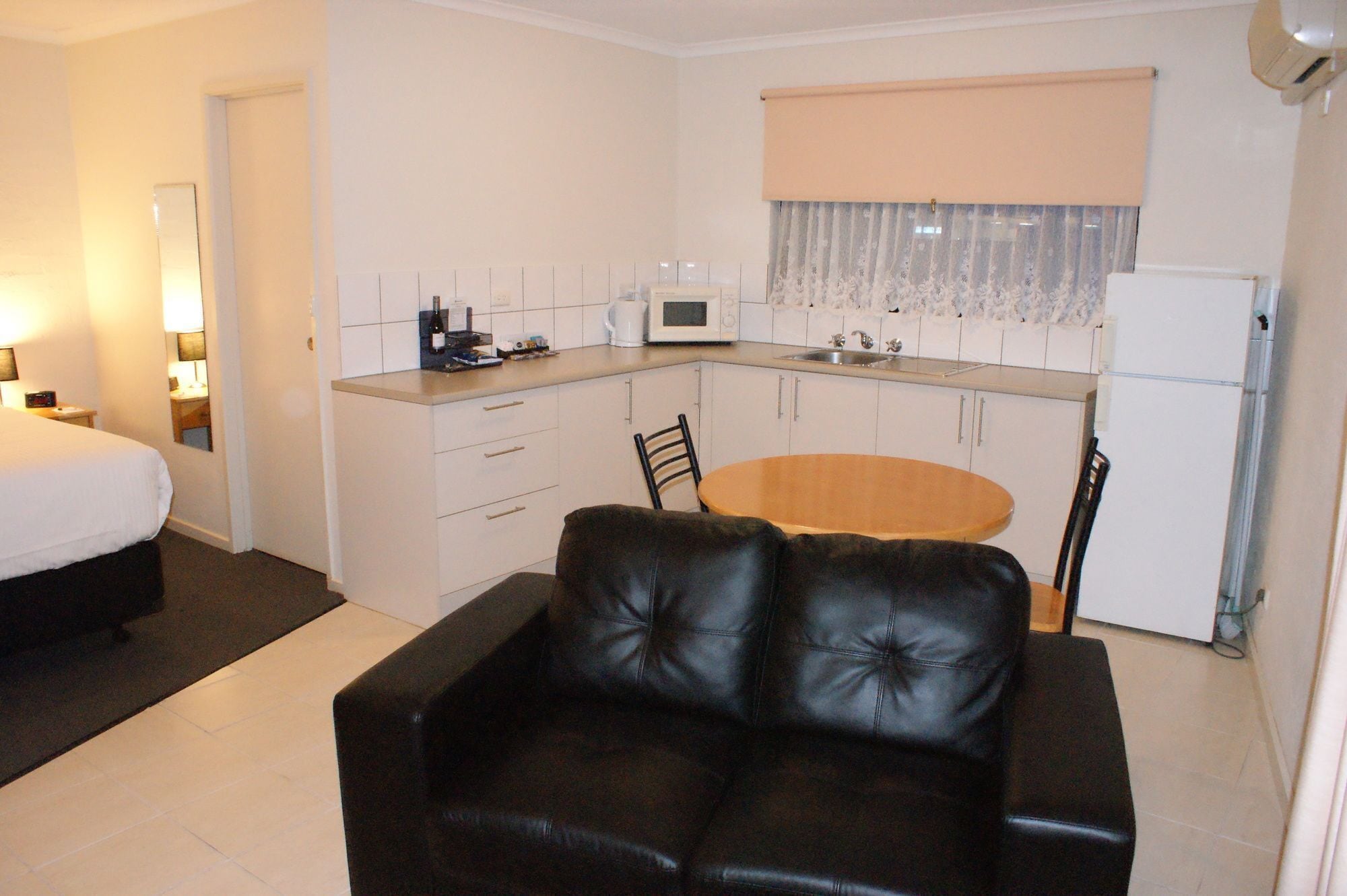 A compact hotel room with a kitchenette, a bed, a black leather sofa, and a dining table.