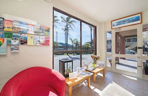 A room with a large window, a red chair, a wooden table with brochures, and a bulletin board with travel posters. - Apollo Bay Motel & Apartments (Apollo Bay)