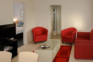 A modern living room with a flat-screen TV, two red armchairs, a white sofa, and a glass coffee table. - Apollo Bay Motel & Apartments (Apollo Bay)