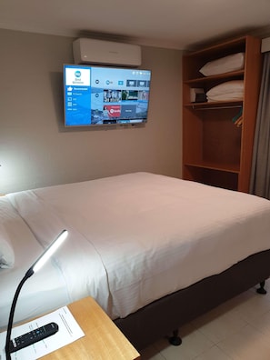 Una camera d'hotel con un letto, un comodino con telecomando, una TV a muro che mostra l'interfaccia di un servizio di streaming e un armadio in legno.