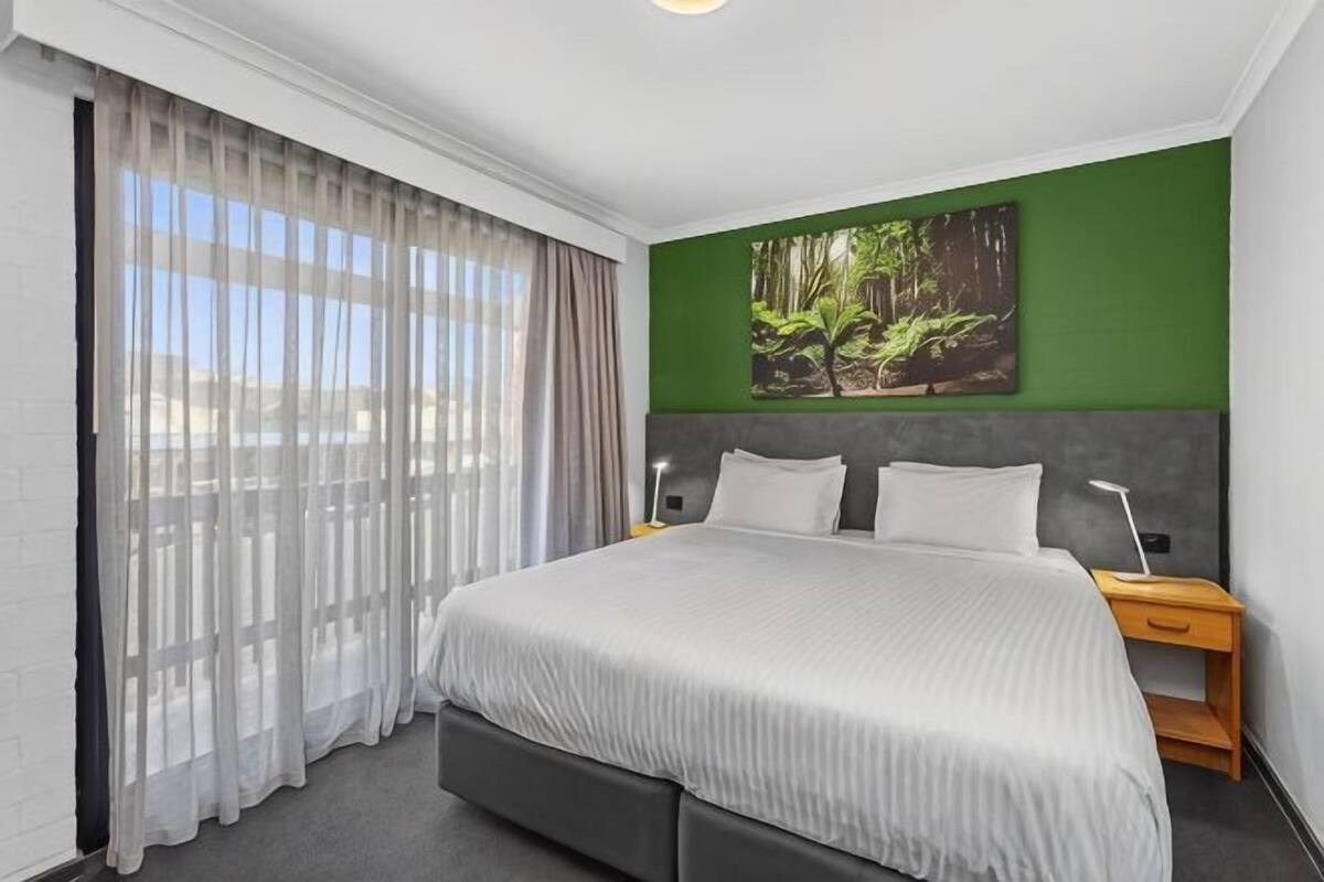 A modern hotel room with a large bed, a nightstand, a green accent wall, and a view through sheer curtains.