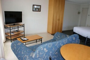 A living room with a sofa, a coffee table, a TV on a stand, and a bed. - Apollo Bay Motel & Apartments (Apollo Bay)