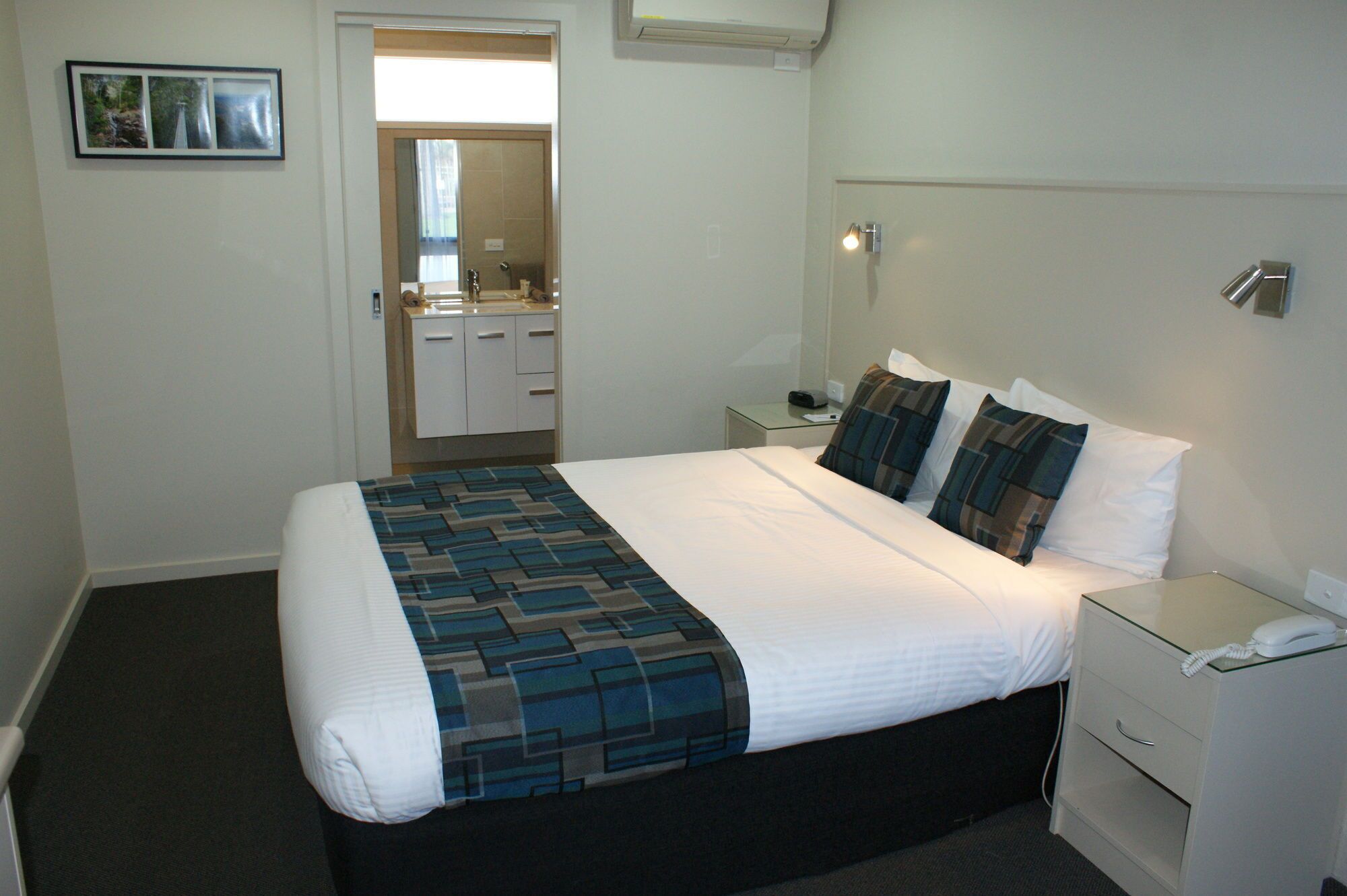 A hotel room with a bed, bedside tables, a nightstand, a mirror, and a door leading to a bathroom.