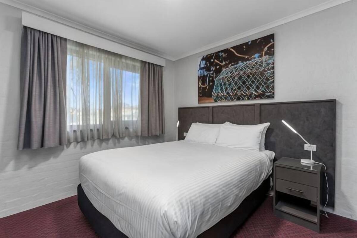 A modern hotel room with a large bed, a nightstand, a desk lamp, and a window with curtains.
