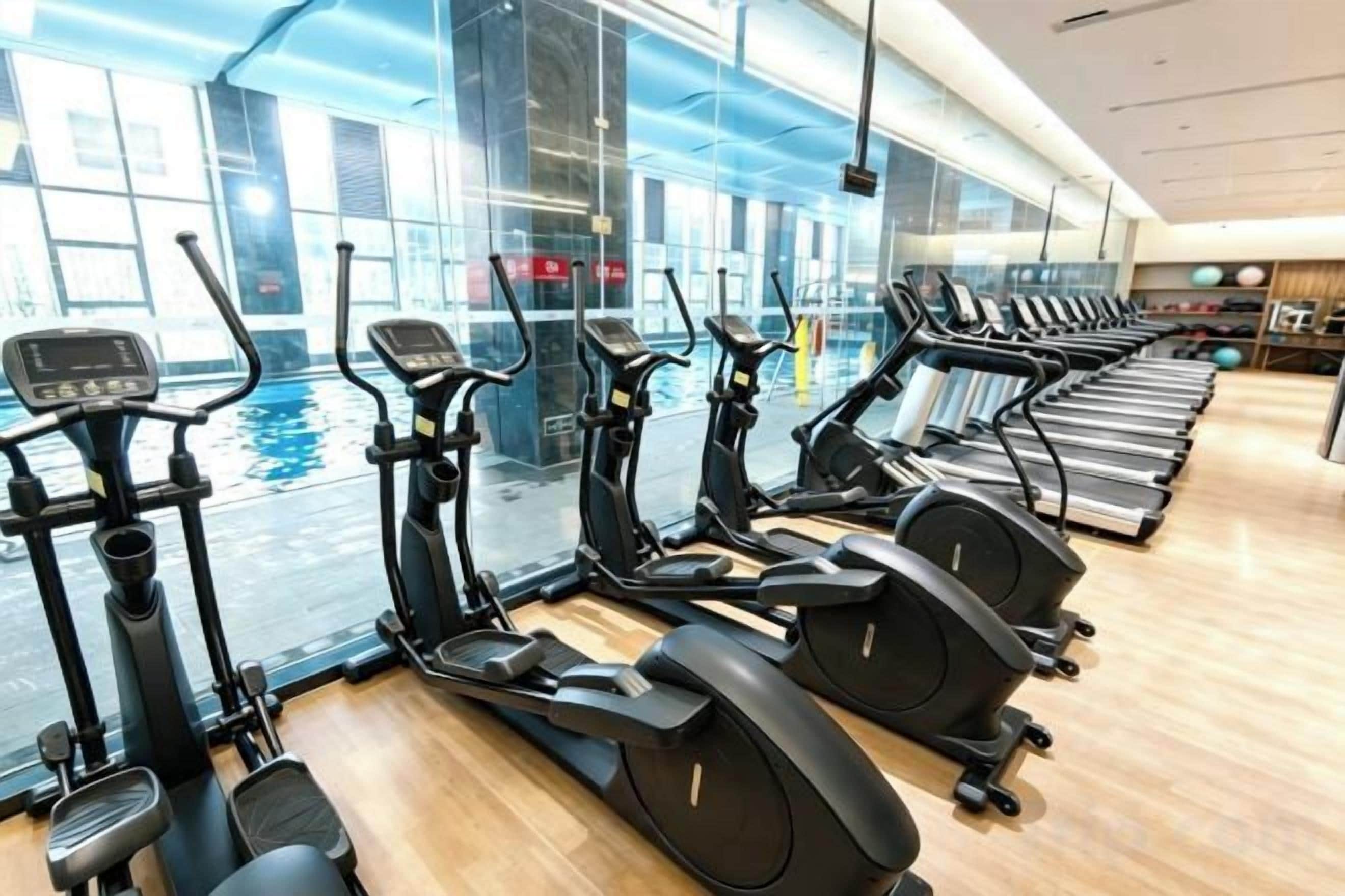 Une salle de sport avec plusieurs tapis de course alignés en rangées, une grande fenêtre donnant sur une piscine et un plancher en bois.