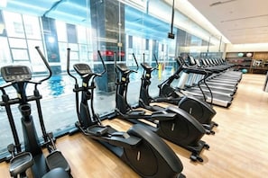 Een fitnessruimte met meerdere loopbanden opgesteld in rijen, een groot raam met uitzicht op een zwembad en een houten vloer.