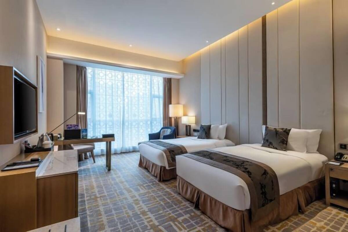 A hotel room with two beds, a desk, a TV, and a large window with curtains.
