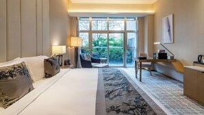 Habitación de hotel con cama, escritorio, silla, lámpara y vista a zonas verdes.