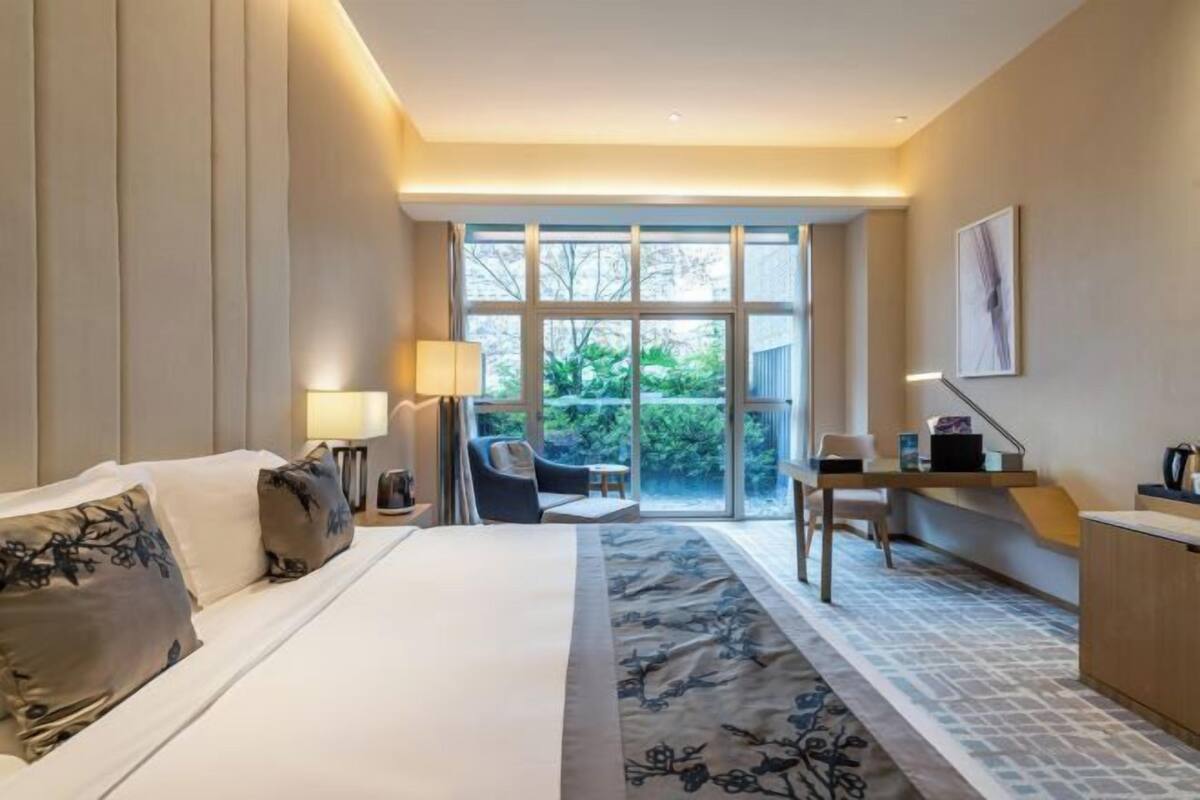 Habitación de hotel con cama, escritorio, silla, lámpara y vista a zonas verdes.