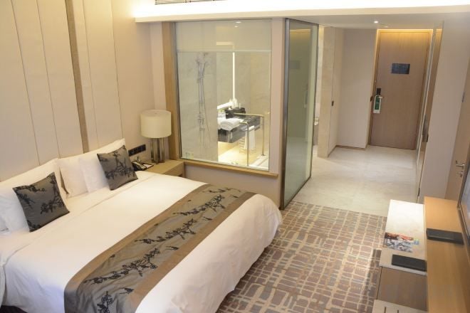A hotel room with a bed, a nightstand, a mirror, and a bathroom visible through a glass door.
