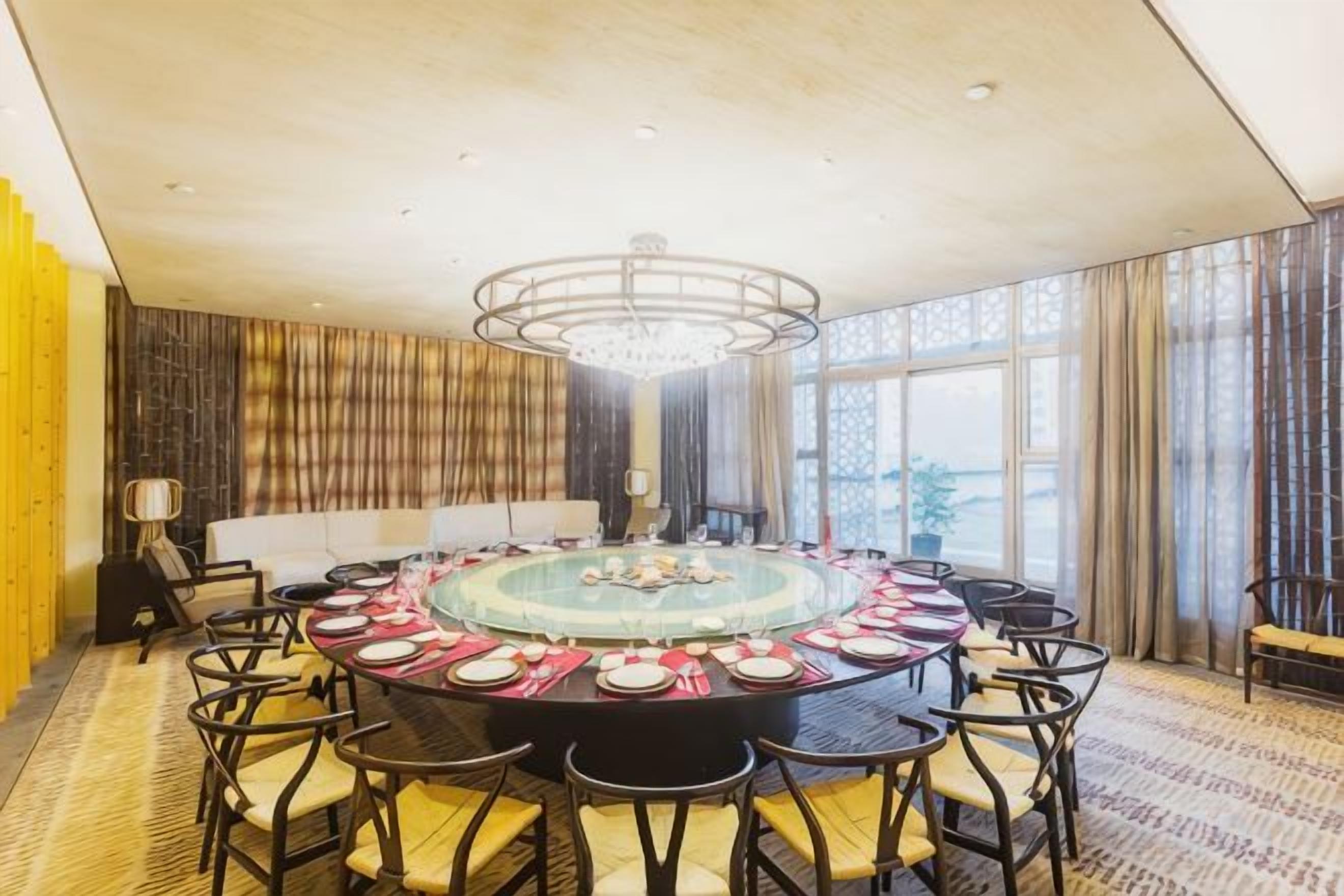 A round table with chairs, a chandelier, and a view of the outdoors through large windows.
