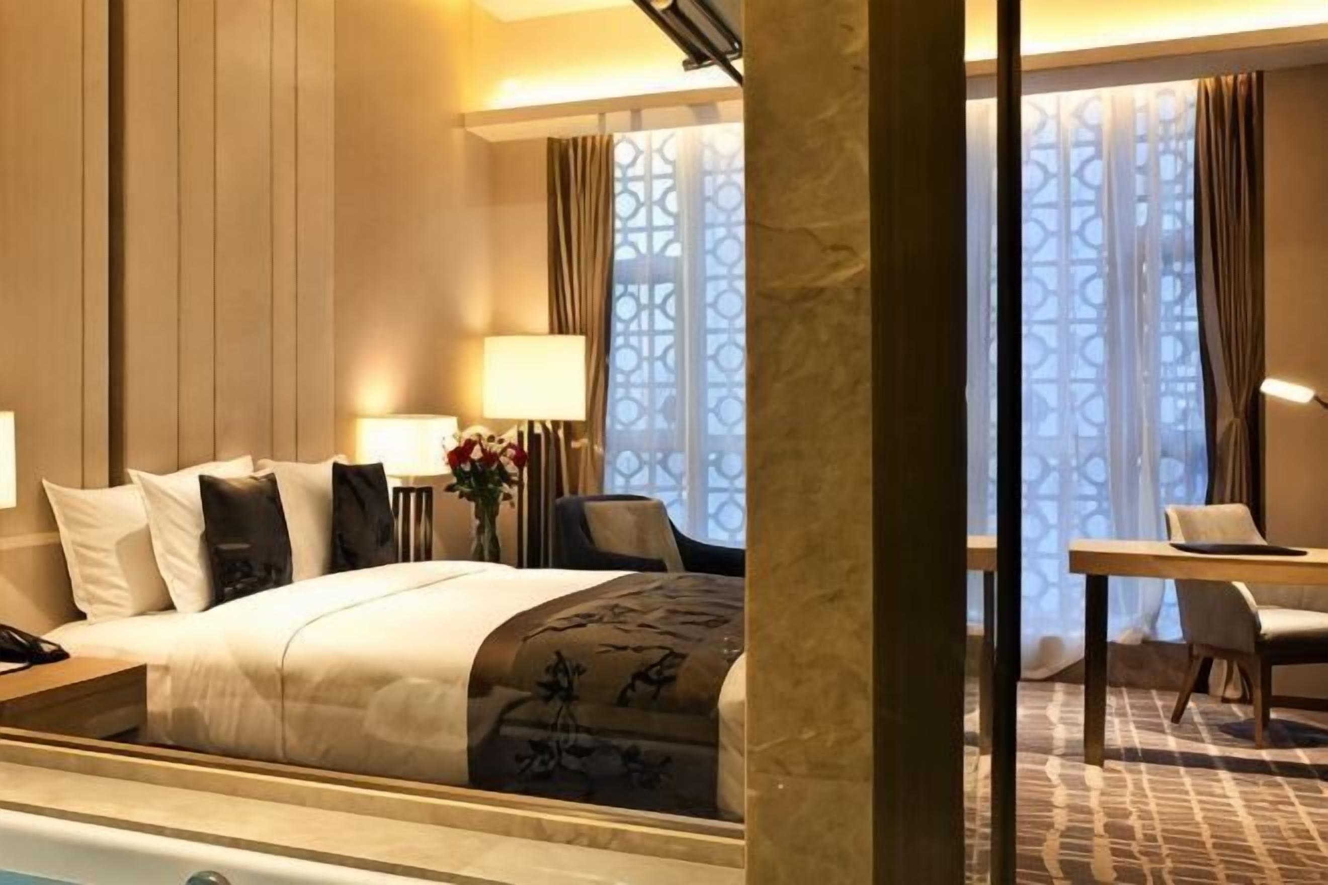 Habitación de hotel con cama, mesita de noche, lámpara, silla y vistas a través de una ventana con cortinas.