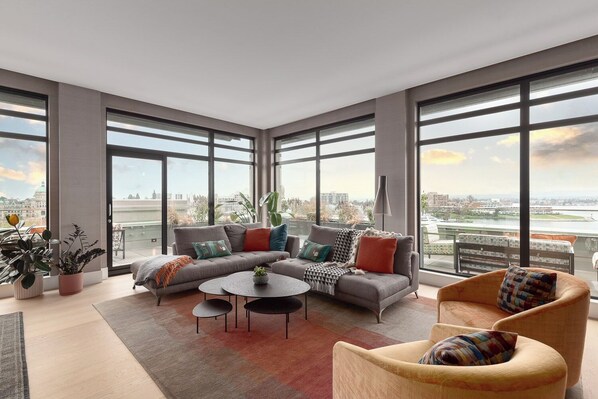 A modern living room with a sectional sofa, two armchairs, and a coffee table. Large windows offer a city view. - Harbour Vista (Victoria)